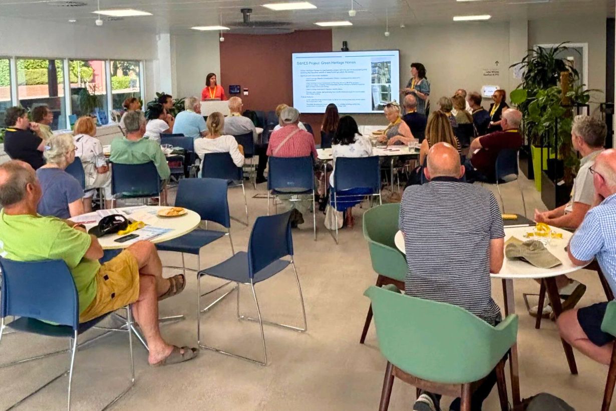 Photograph of a community meeting in a modern room with multiple round tables and chairs arranged for attendees. Two presenters stand near a screen displaying a slide, while around 25 people listen, highlighting an informative group discussion or workshop setting.