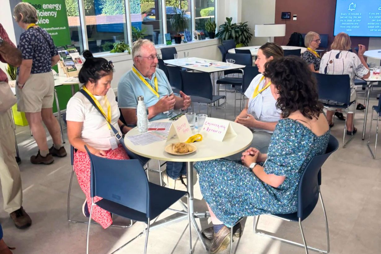 Photograph of a group discussion in a bright, modern room with four people seated around a round table, each wearing yellow lanyards. Table holds name cards and snacks, with informational posters and other participants visible in background, suggesting a workshop or community event focused on energy efficiency.