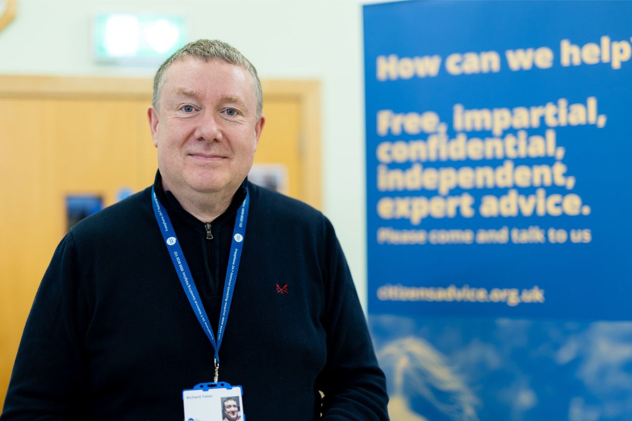 Photograph of a man wearing a black sweater and blue lanyard standing indoors next to a blue banner. Banner text promotes free, impartial, confidential, independent expert advice with a website link for citizens advice.
