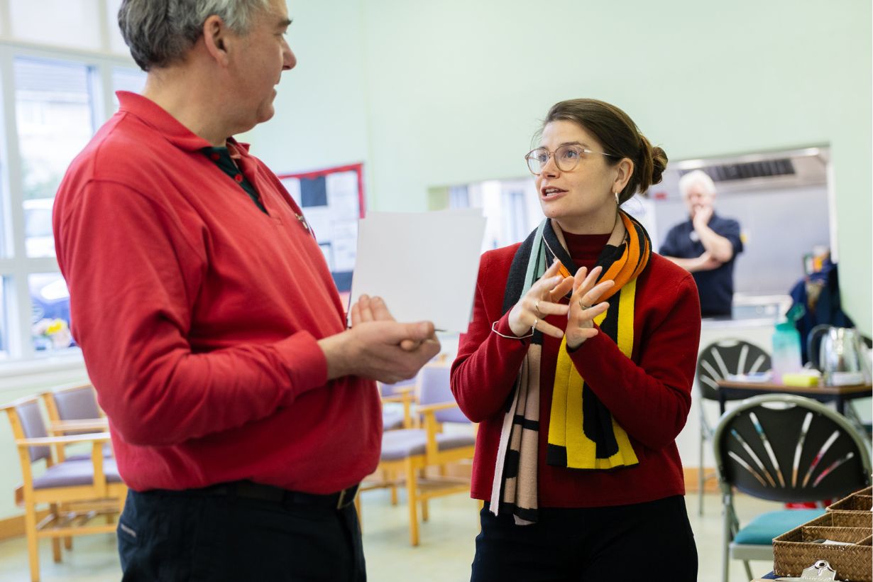 Photograph of two people engaged in conversation inside a room with chairs and tables, with a third person blurred in the background. The woman wears glasses and a colourful scarf, while the man holds a sheet of paper and wears a red shirt.