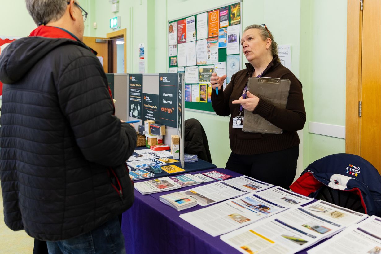 Photograph of an indoor information stall where a woman holding a clipboard speaks to a man in a black jacket. Table covered with pamphlets, brochures, and informational posters about energy related support and services, set against a bulletin board with various notices.