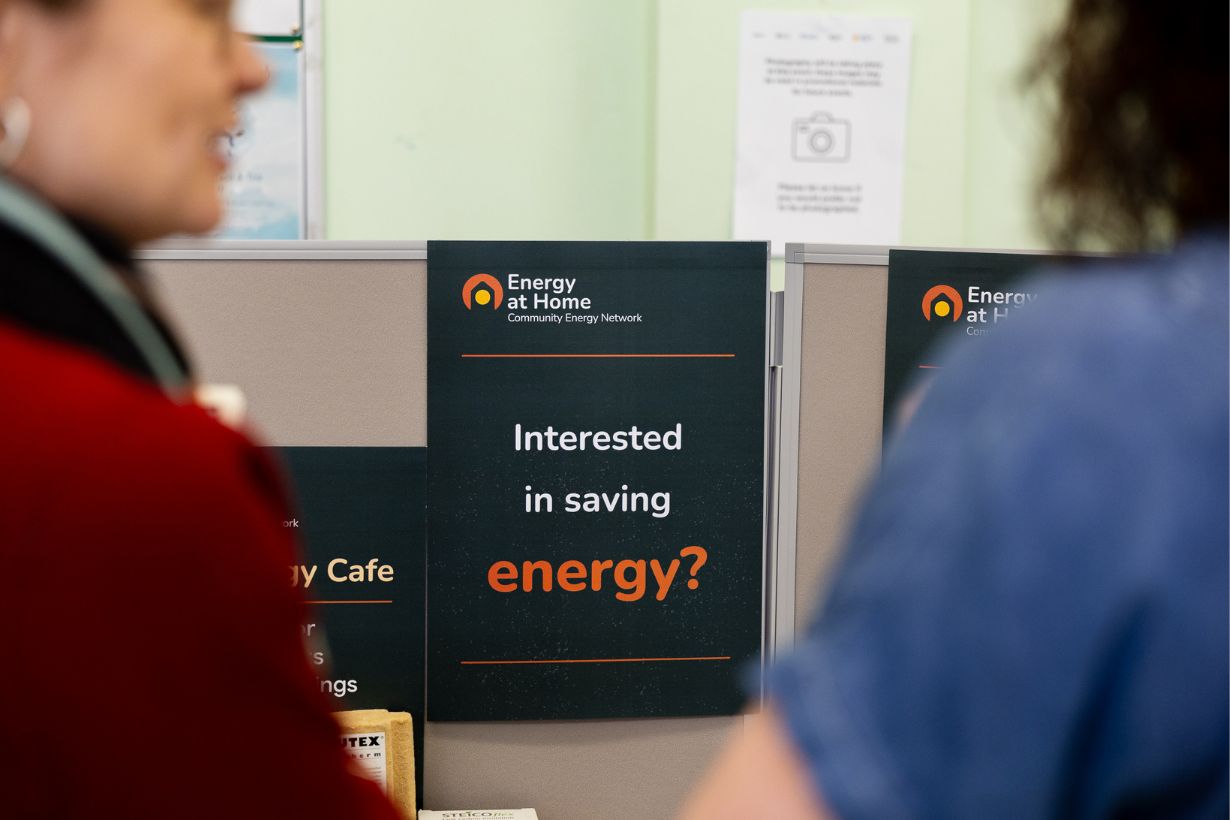 Photograph showing a promotional sign for Energy at Home ork with text "Interested in saving energy?" positioned between two blurred individuals. Sign features a black background with white and orange text, highlighting an energy-saving initiative.