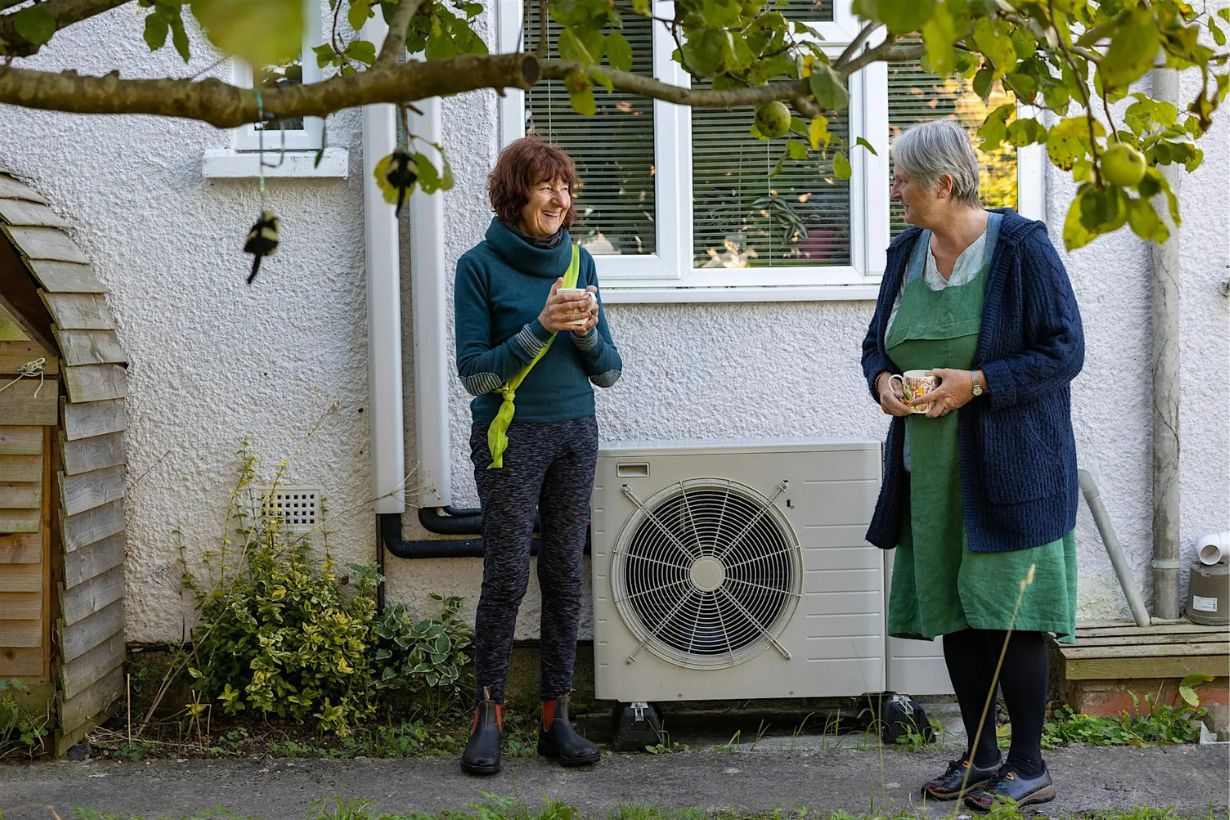 Two women beside a house, discussing an outdoor air-source heat pump.