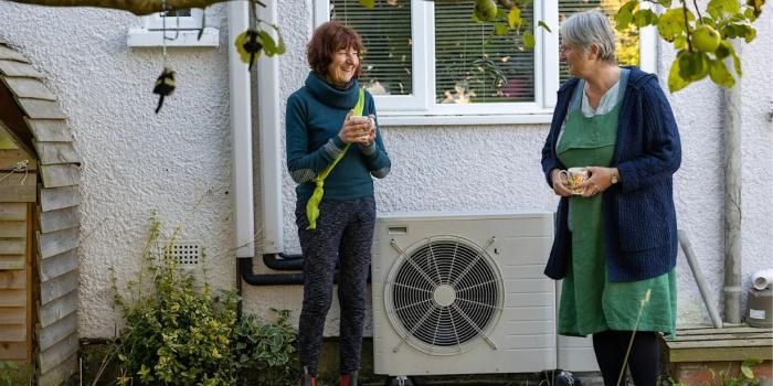 Two women beside a house, discussing an outdoor air-source heat pump.