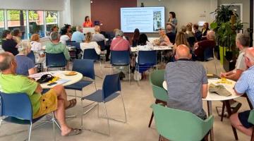 Photograph of a community meeting in a modern room with multiple round tables and chairs arranged for attendees. Two presenters stand near a screen displaying a slide, while around 25 people listen, highlighting an informative group discussion or workshop setting.