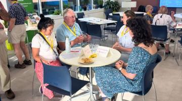 Photograph of a group discussion in a bright, modern room with four people seated around a round table, each wearing yellow lanyards. Table holds name cards and snacks, with informational posters and other participants visible in background, suggesting a workshop or community event focused on energy efficiency.