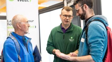 Three men engaged in conversation at a Retrofit West event, with one man wearing a green Retrofit West branded jacket. A banner in background promotes energy-efficient home improvements, highlighting the context of sustainable housing and retrofit solutions.