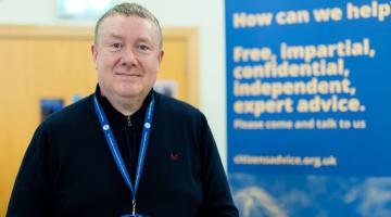 Photograph of a man wearing a black sweater and blue lanyard standing indoors next to a blue banner. Banner text promotes free, impartial, confidential, independent expert advice with a website link for citizens advice.