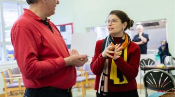 Photograph of two people engaged in conversation inside a room with chairs and tables, with a third person blurred in the background. The woman wears glasses and a colourful scarf, while the man holds a sheet of paper and wears a red shirt.