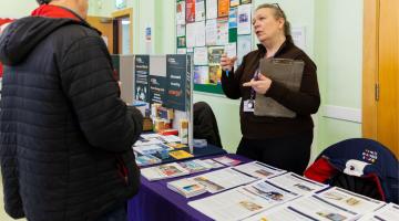 Photograph of an indoor information stall where a woman holding a clipboard speaks to a man in a black jacket. Table covered with pamphlets, brochures, and informational posters about energy related support and services, set against a bulletin board with various notices.