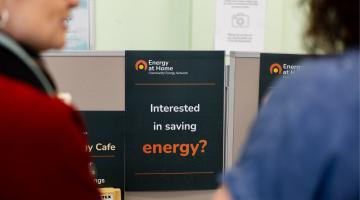 Photograph showing a promotional sign for Energy at Home ork with text "Interested in saving energy?" positioned between two blurred individuals. Sign features a black background with white and orange text, highlighting an energy-saving initiative.