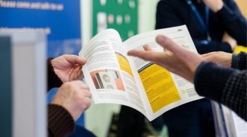 Photograph showing two people holding and discussing an open factsheet with text and images, including a section highlighted in yellow. Background features a blurred person and a blue banner with partially visible text, suggesting an informational or advisory setting.
