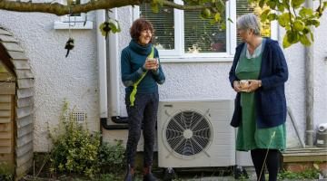 Two women beside a house, discussing an outdoor air-source heat pump.